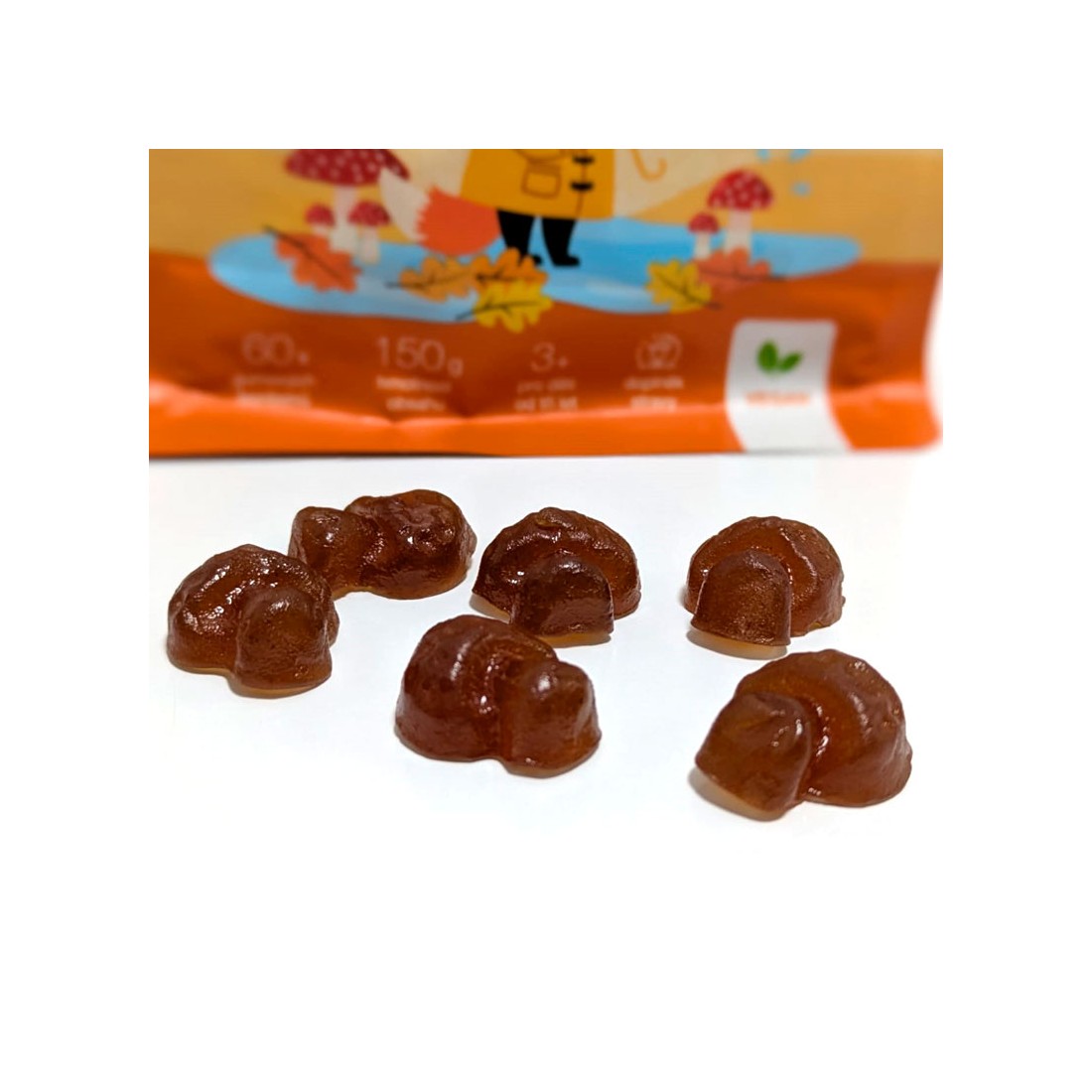 Gominolas marrones con forma de seta frente a su envase naranja de MycoGummies de 150g con ilustraciones de bosque.